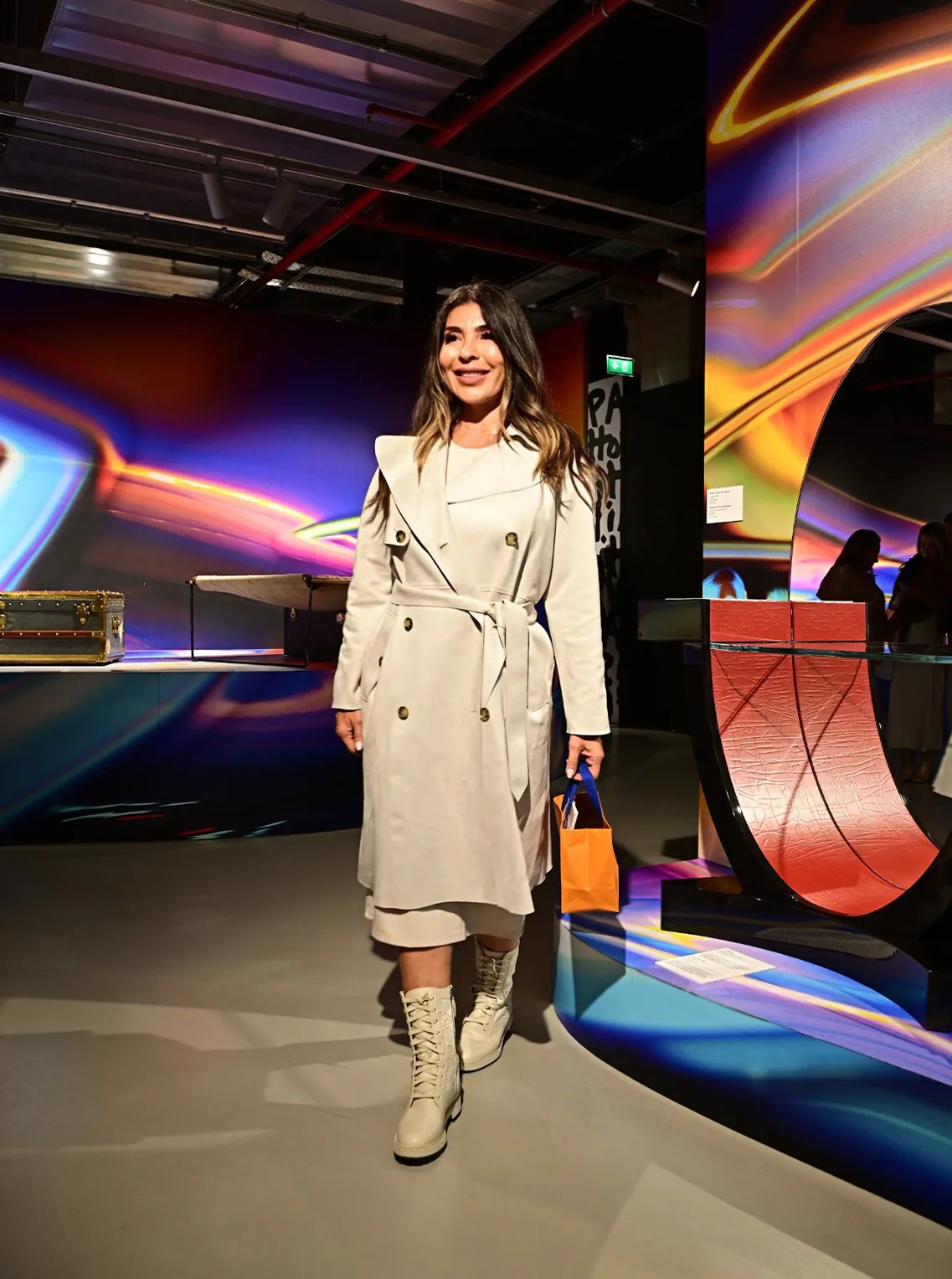 Woman in a belted beige trench coat and white boots at a colorful art installation