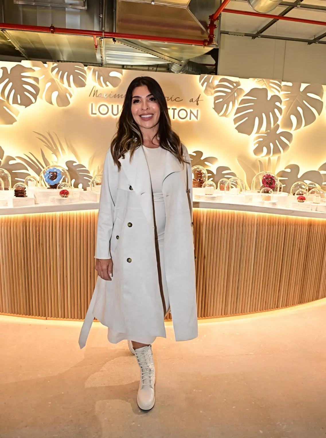 Woman in a long white coat at a Louis Vuitton event with a palm-leaf backdrop